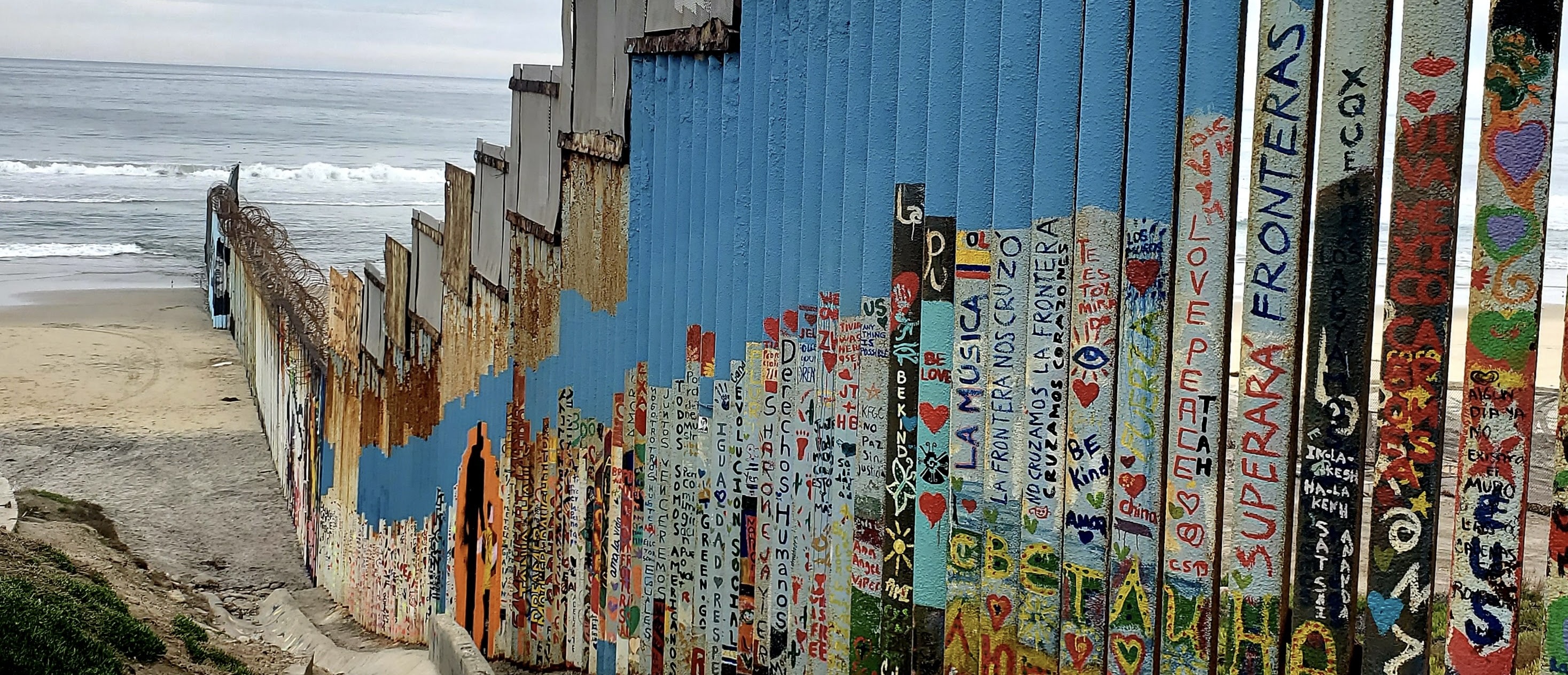 The US-Mexico border wall at Playas de Tijuana covered in colorful murals and messages of hope, extending into the Pacific Ocean